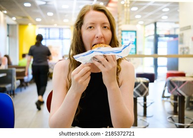 Woman With Burger In A Fast Food Restaurant. Woman Opening Wrapping Paper, Holding, Biting The Burger And Savouring Food. Fast Snack On The Go