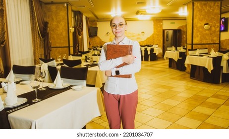 Woman Business Owner. Portrait Of A Female Waitress In A White Shirt Holding A Restaurant Menu With Both Hands. Girl Entrepreneur Or Cafe Employee Posing In Restaurant Coffee Shop Interior.