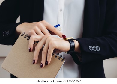 Woman in a business suit and dark red manicure holding a notebook and looks at his watch close-up