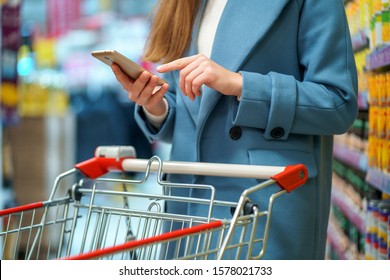 Woman Buyer With Cart In The Store Aisle With Grocery List On Smartphone During Shopping Food 