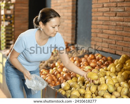 Woman buyer collects selects products in store according to list for weekly purchase. Female customer made beeline for fresh produce section, pick potato and puts it in bag