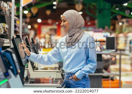 A woman buyer in a supermarket in a hijab pays for goods at a self-service checkout, convenient service for customers.