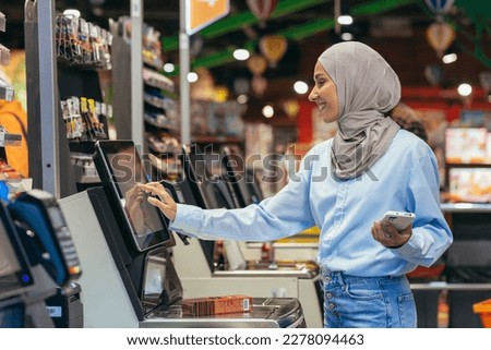 A woman buyer in a supermarket in a hijab pays for goods at a self-service checkout, convenient service for customers.