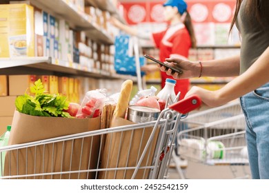 Woman buying groceries at the supermarket, she is pushing a shopping cart and using her smartphone, unrecognizable people - Powered by Shutterstock