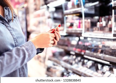 Woman Buying Make Up At Cosmetics Section In Store. Customer Shopping Beauty Products. Choosing Red Lipstick From Shelf And Selection. Using Tester. Makeup Business Concept. Supermarket Or Drugstore.