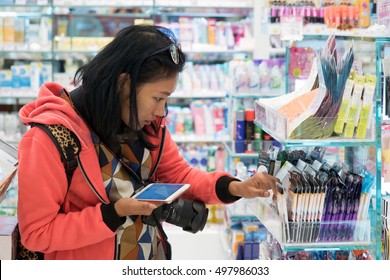 Woman buys cosmetics in the store. Asian girl is looking at items in  shop. Consumers buy with help of smart-phone in hand. People choose cosmetics products with phone assistance. Check price at shop.