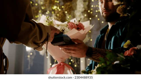 Woman buys spring bouquet in flower shop and pays for purchase with contactless payment using smartphone. Male florist sells fresh flowers to female customer. Floristry and entrepreneurship concept. - Powered by Shutterstock