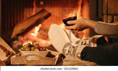 Woman By The Fireplace. Woman In Woollen Socks Taking A Glass Of Red Wine Relaxing By The Cozy Fireplace. Snug Winter Evening.