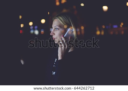 Woman calling with cell telephone and using city computer for touristic information while standing at night on the street, girl touching digital display on bus stop during mobile phone conversation 