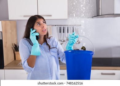 Woman Calling Plumber While Collecting Water Leaking From Ceiling Using Bucket