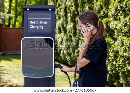 Woman Calling While Using EV Charger. Young woman talks on a smartphone while holding an electric vehicle charging cable at an outdoor destination charging station.