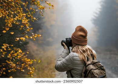 Woman with camera taking picture of autumn leaf. Tourist hiking in misty forest. Landscape photographer with backpack enjoying nature in fall season - Powered by Shutterstock