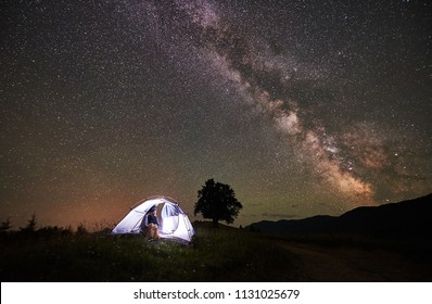 Woman Camper Enjoying At Night Camping In The Mountains Under Amazing Night Sky Full Of Stars And Milky Way. Girl Sitting Inside Illuminated Tent And Looking At Sky Full Of Stars. Astrophotography