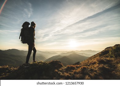 The woman with a camping backpack standing on a rock with a picturesque sunset