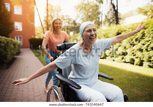 A\
woman with cancer is sitting in a wheelchair. She walks on the\
street with her daughter and they fool around. They are fun and\
they laugh. They walk in the courtyard of the\
clinic.