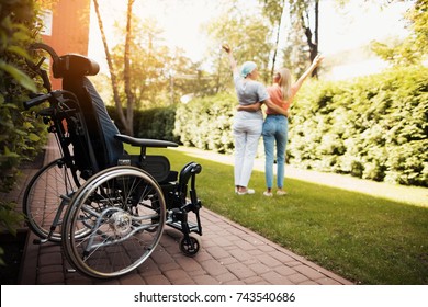 A woman with cancer stands up with her daughter. They embrace. Nearby is a wheelchair woman. They are in the courtyard of a modern clinic. - Powered by Shutterstock