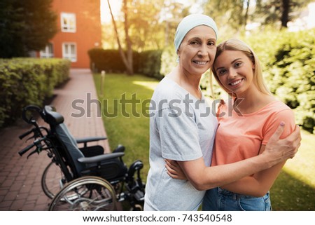 A woman with cancer stands up with her daughter. They embrace. Nearby is a wheelchair woman. They are in the courtyard of a modern clinic.
