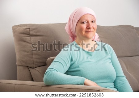 Woman with cancer and wearing a pink scarf smiling sitting on a sofa at home. Smiling woman suffering from cancer sitting after taking chemotherapy sessions.