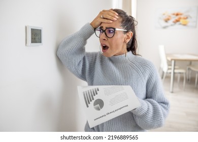 Woman Cannot Believe Her Own Eyes The Sum On Her Energy Bill Looking Shocked At The Thermostat On The Wall Next To Her.