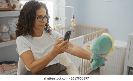 Woman capturing moments on a phone while seated in a cozy bedroom with a cradle, surrounded by soft toys, showcasing a peaceful indoor setting.
