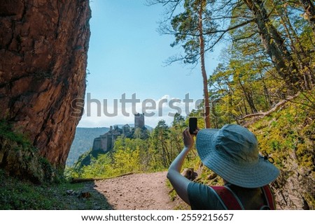 A woman capturing the view of Chateau de Fleckenstein in Alsace, surrounded by dense greenery and rocky landscapes under a bright blue sky