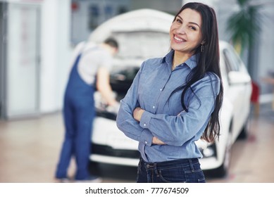 Woman At A Car Garage Getting Mechanical Service. The Mechanic Works Under The Hood Of The Car.