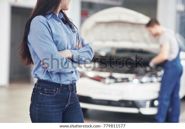 Woman at a car garage getting\
mechanical service. The mechanic works under the hood of the\
car.