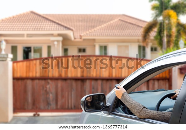 Woman in car,\
hand using remote control to open the wooden automatic gate with\
modern  home blurred background when  arrived home. Home remote\
control and auto door in\
concept.