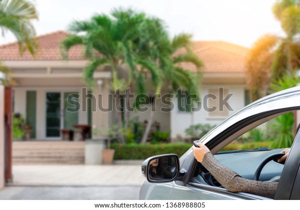 Woman in car, hand using remote\
control to open the auto gate with modern home blurred background\
when  arrived home. Home remote control and automatic door in\
concept
