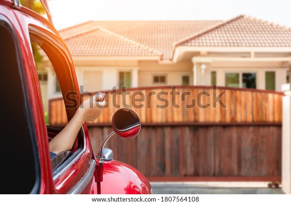 Woman in car, hand using\
remote control to open the automatic gate with modern home blurred\
background. The electric door and home remote control auto door in\
concept.