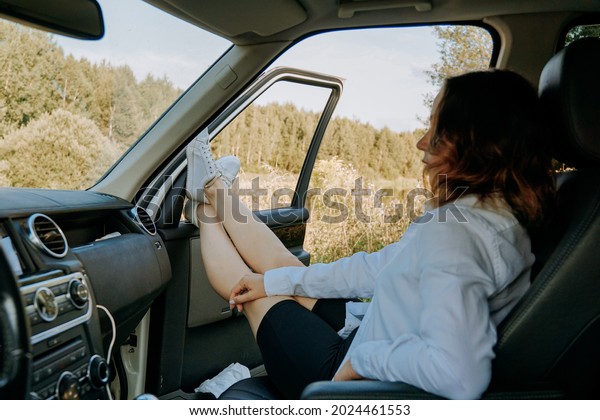 The woman in the car in the passenger seat.\
Feet out the window. Landscape view through the window. A trip to\
nature, rest outside the\
city.