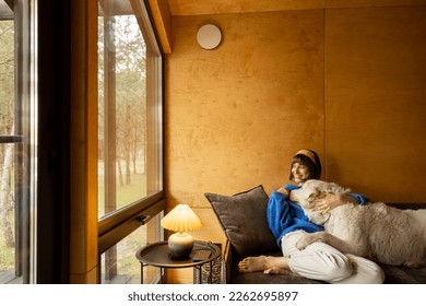 Woman cares her dog while sitting on a couch by the window, spending leisure time together in wooden house on nature - Powered by Shutterstock