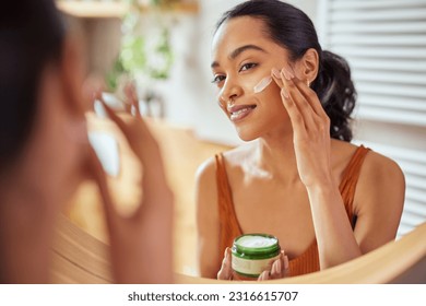 Woman caring of her beautiful skin on the face standing near mirror in the bathroom. Mexican woman applying moisturizer on her face at home. Multiethnic girl holding little green jar of bio skin cream