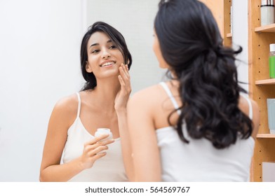Woman caring of her beautiful skin on the face standing near mirror in the bathroom. Young woman applying moisturizer on her face. Smiling girl holding little jar of skin cream and applying lotion.