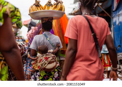 Woman carries baby on back in the traditional manner while carrying bananas on her head. In the market in Cotonou, Benin. 