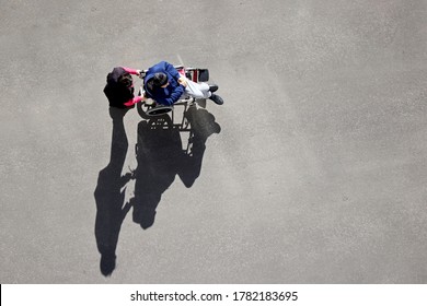 Woman Carries A Disabled Person In A Wheelchair On A City Street, Top View. Nurse And Elderly Man, Care For Handicapped People