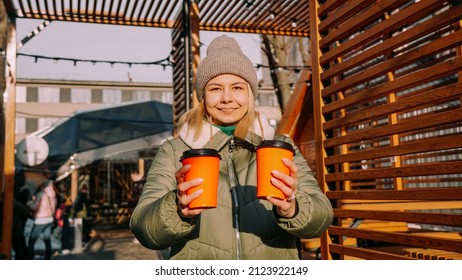 Woman Carries Two Cups Of Coffee Or Mulled Wine At Outside Food Court. Street Food Festival, Place To Relax With Friends. She Holds The Cups Forward