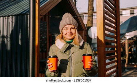 Woman Carries Two Cups Of Coffee Or Mulled Wine At The City Outside Food Court. She Looks At The Camera. Street Food Festival, Place To Relax And Eat