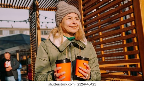 Woman Carries Two Cups Of Mulled Wine To Her Friend At The City Outside Food Court. Street Food Festival, Place To Meet Friends