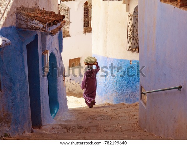 A woman carring the bread on her\
head and walking down the blue-white streets in Chefchaouen - the\
blue city Morocco - amazing palette of blue and white\
buildings