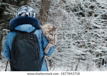 Woman carrying a cat outside, in the winter forest. Fluffy, gray cat sitting on the shoulder of the hostess and looking to the side.