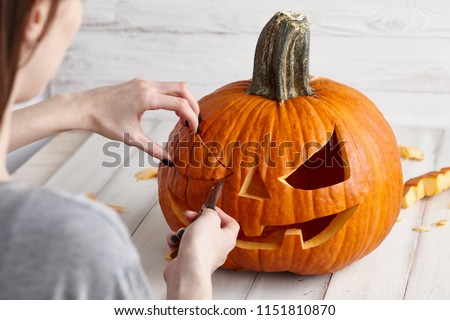 Woman carving big orange pumpkin into jack-o-lantern for Halloween holiday decoration on white wooden planks, close up view