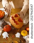 Woman carving halloween pumpkin on table with ghosts and spiders. Carving jack o lantern, halloween preparation. Making halloween decorations