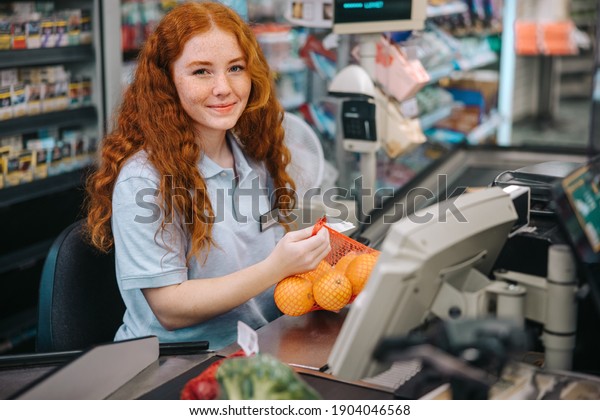 Woman cashier sitting behind checkout  and\
working in supermarket. Female checkout counter clerk scanning\
grocery products and looking at\
camera.