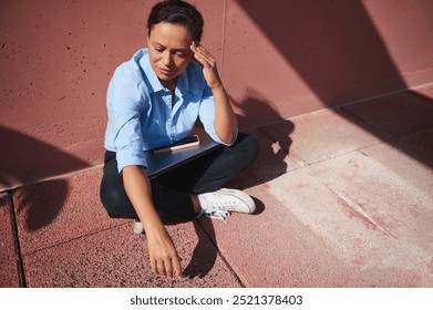 Woman in casual attire sitting outside on a sunny day, contemplating. Shadows and textures create a reflective mood. - Powered by Shutterstock