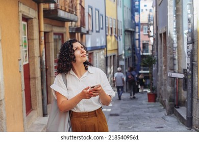 Woman in casual clothes looking up, holding a smartphone, lost on a small European street