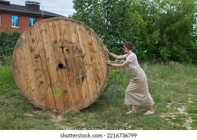 A woman in casual clothes rolls a huge wooden spool of wire