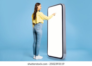 Woman in casual outfit standing in front of large smartphone, touching empty display, using newest mobile app or website, blue background, mockup