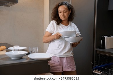 Woman in casual wear loading ceramic plates into dishwasher in a modern kitchen. She is organizing dishes while preparing a meal, creating a tidy and clean environment. - Powered by Shutterstock