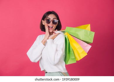 Woman in casual white sweater and sunglasses on pink red background holding shopping bags shocked amazed excited  isolated copy space
